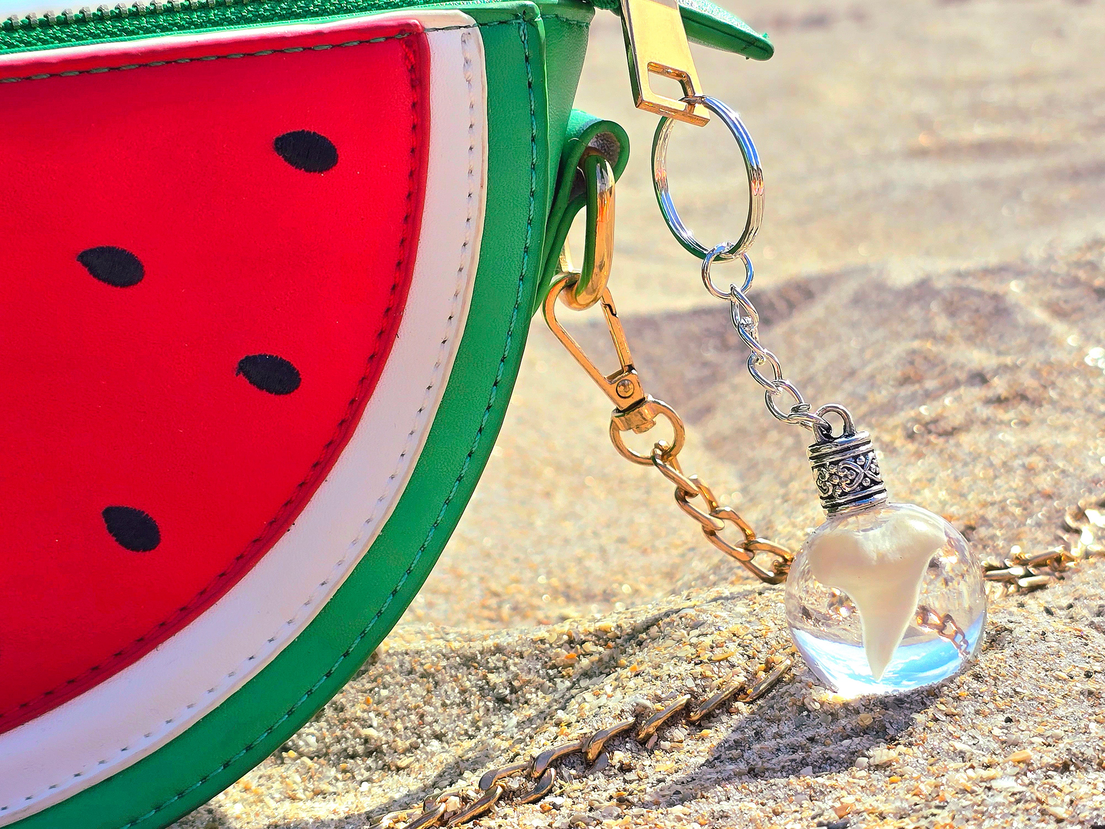 Clear resin sphere keychain holding a real mako shark tooth resting on sand beside a red watermelon purse