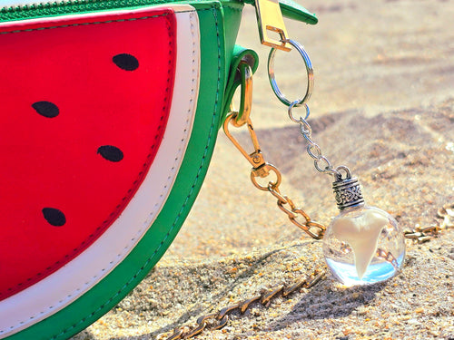 Clear resin sphere keychain holding a real mako shark tooth resting on sand beside a red watermelon purse