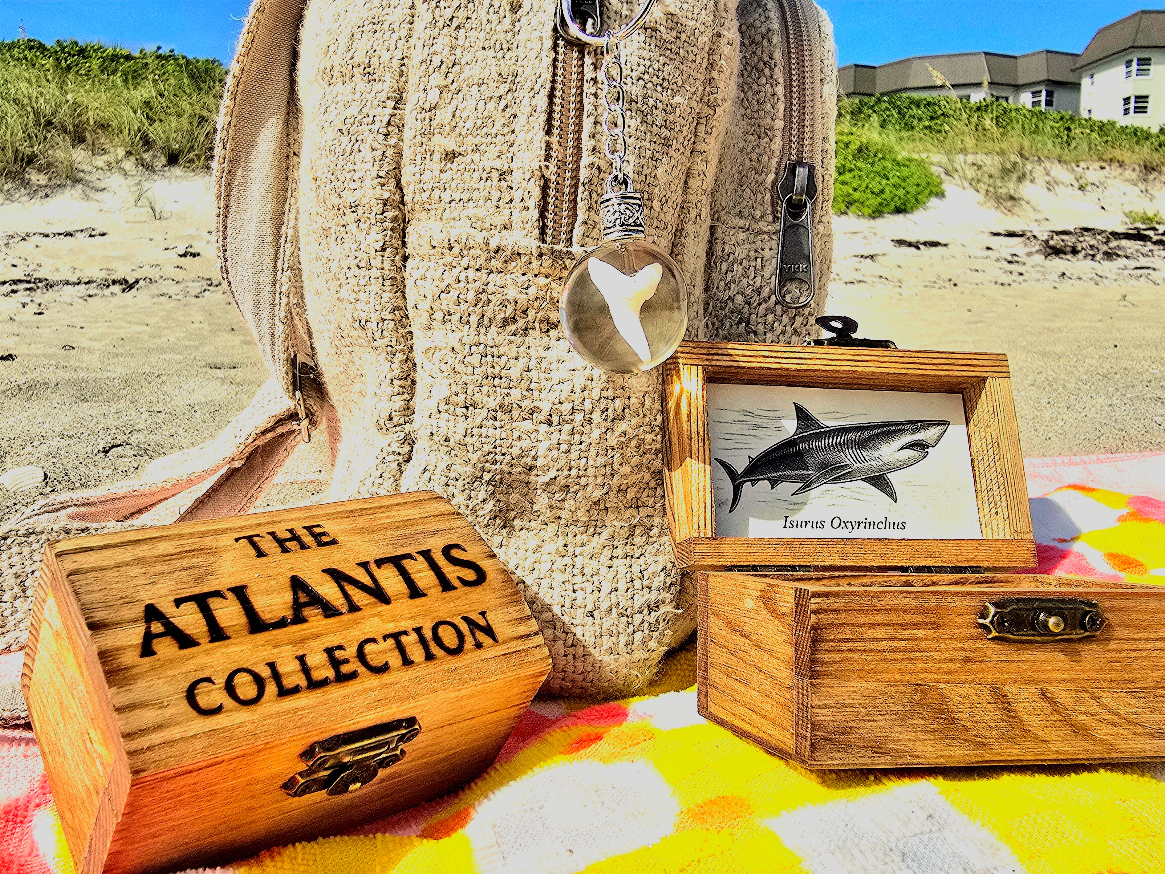 Mako shark tooth keychain hanging from a straw beach bag beside open and closed Atlantis Collection wooden treasure chest boxes