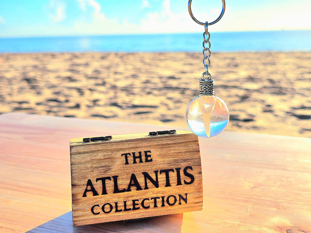Mako shark tooth resin keychain held above an engraved Atlantis Collection wooden box on a sunlit beach