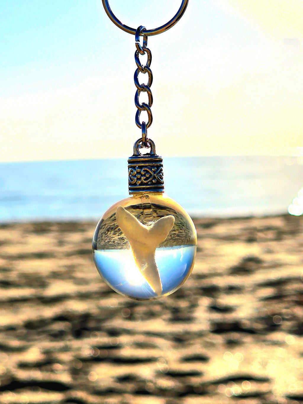 Mako shark tooth in a clear resin sphere keychain held against a golden sandy beach at sunset