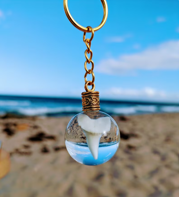 Mako shark tooth resin sphere keychain with a gold chain held against a blue-sky beach